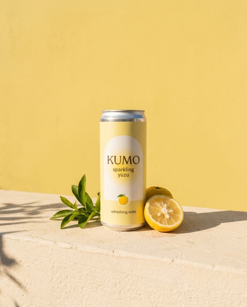 The KUMO can on a cream stone ledge against a yuzu-yellow wall, with yuzus and a sprig of leaves